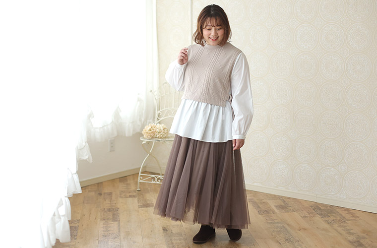 白色のシャツにベージュのショート丈ニットを重ねたようなドッキングトップスを着て、ブラウンのチュールスカートを履く女性