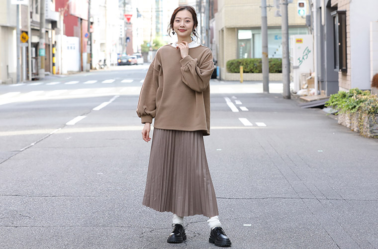 白色のブラウスにブラウンのトップスを重ね着して、同系色のプリーツスカートを履く女性