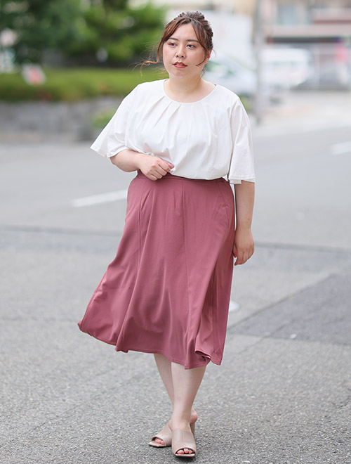 白色のフレアー袖ブラウスを着て、くすみピンク色のAラインスカートを履く女性