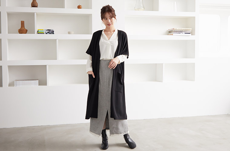 グレーの前スリットロングスカートに白シャツを合わせ、黒の半袖ジレを羽織る女性