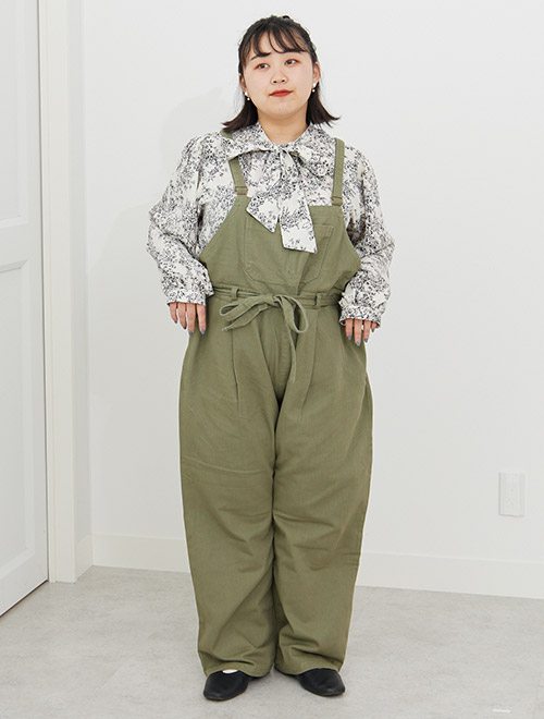 オフホワイトに黒色で柄を描いたリボンタイブラウスを着て、カーキのサロペットを履く女性