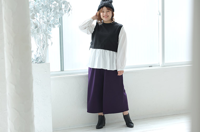 白色のシャツに黒色のショート丈ニットを重ね着したようなトップスを着て、紫色のワイドパンツを履く女性