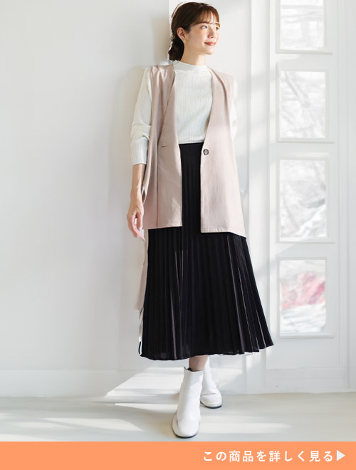 ブラックのアコーディオンスカートとホワイトトップスのコーデに、ベージュのジレを羽織る女性