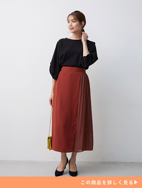 くすみオレンジのサイドプリーツスカートを履き、黒のブラウスをタックインして着る女性