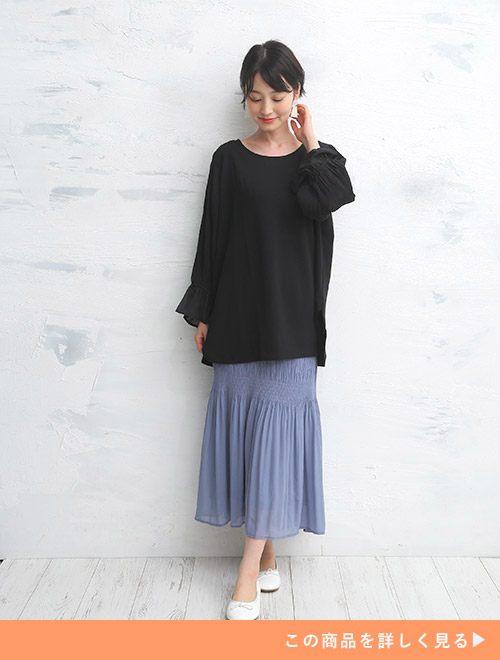 黒のブラウスを着て、ダスティブルーの変形プリーツスカートを履く女性