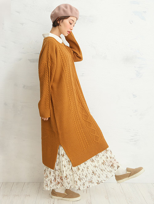 キャメルのロングニットワンピースに、花柄のプリーツスカートを合わせた女性