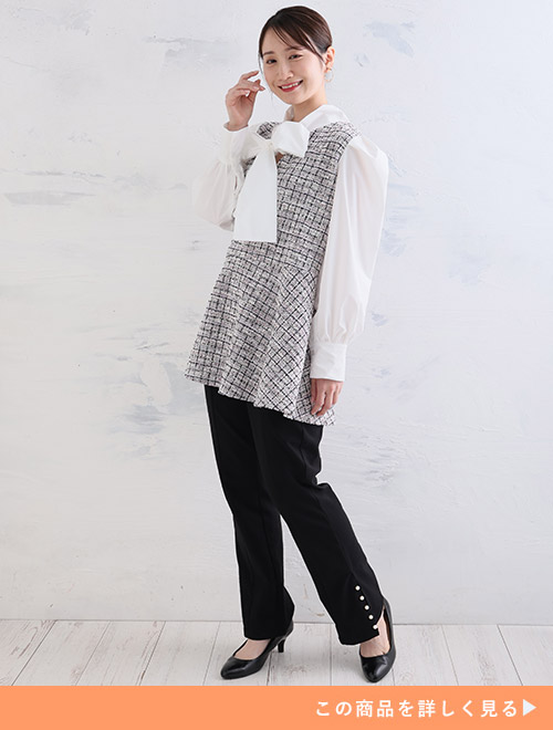 白のリボンタイブラウスにツイードベストを重ねた風デザインのトップスを着て、黒のパンツを履く女性