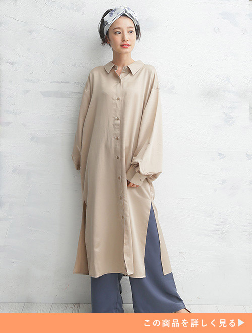 光沢のあるベージュ色のロング丈シャツワンピースを着て、くすみブルーのパンツを履く女性