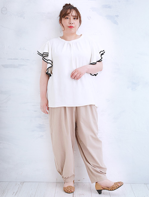 袖口を黒でパイピングしたオフホワイトのフリル袖トップスに、ベージュのパンツを履く女性