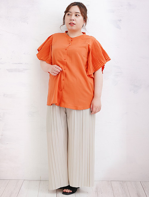 フリル袖部分をプリーツにしたオレンジ色ブラウスに、アイボリーのワイドパンツを履く女性