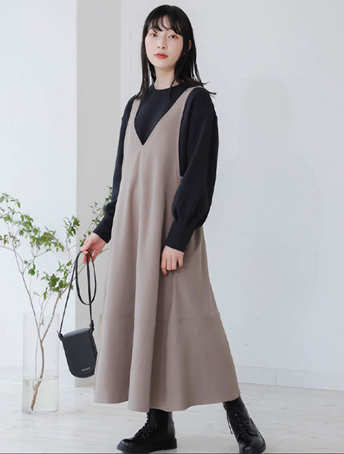モカベージュのVネックジャンパースカートを着て、インナーに黒トップスを合わせる女性