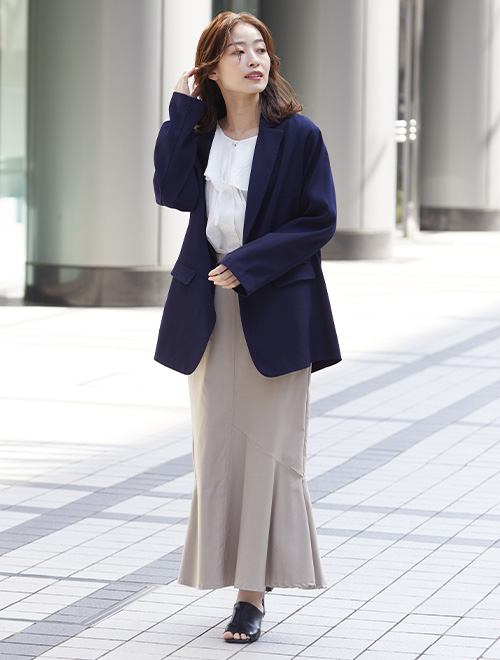 フリルブラウスにネイビーのジャケットを羽織り、グレージュのマーメイドスカートを履く女性
