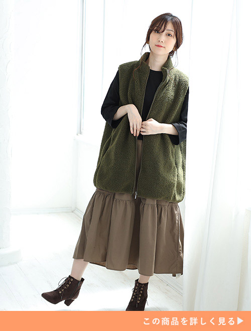 黒トップス×モカ色スカートのドッキングワンピースに、カーキ色のファーベストを羽織る女性