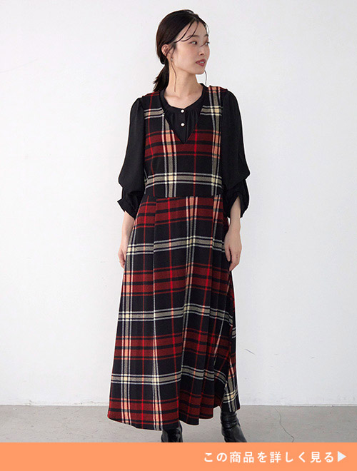 黒ブラウスにオレンジ系のチェック柄ジャンパースカートを重ね着する女性。