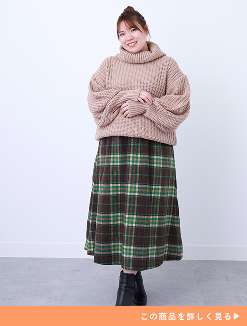 ベージュ色のチャンキーニットトップスを着て、茶×緑系のチェック柄スカートを履く女性