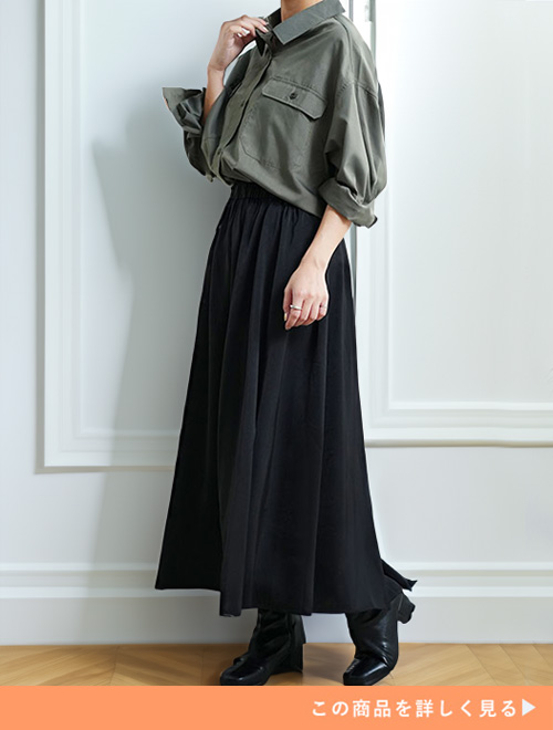 ブラックのサテンフレアスカートとカーキのシャツのコーデにヒールブーツを履く女性