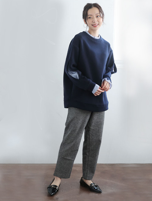 レイヤード風トップスとグレーのボトムスを着た女性が前で手を組んで右側を向いている