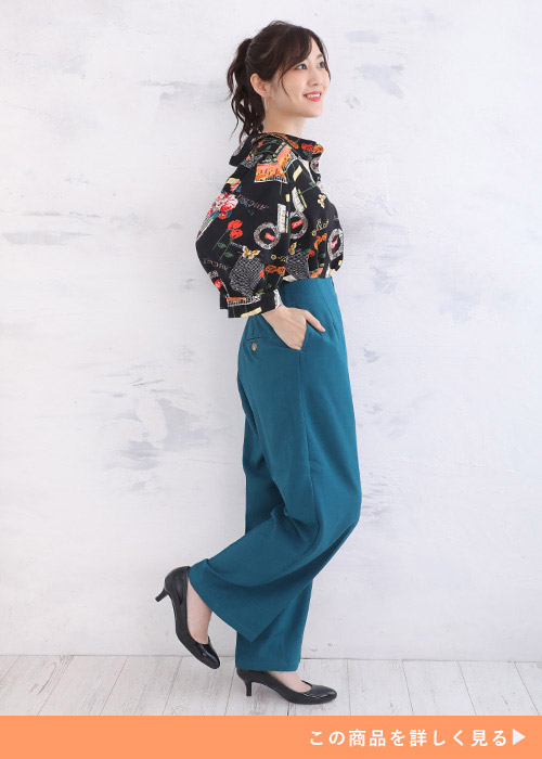 黒地に大胆なレトロ柄が描かれたブラウス×ダックブルーのカラーパンツコーデを着る女性