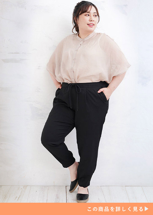 ブラックのパンツにベージュのとろみシャツを着て、右足のかかとを上げて左を向く女性