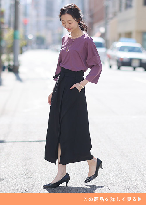 パープルのトップスとブラックのスリットスカートのコーデに黒のヒールパンプスを履く女性