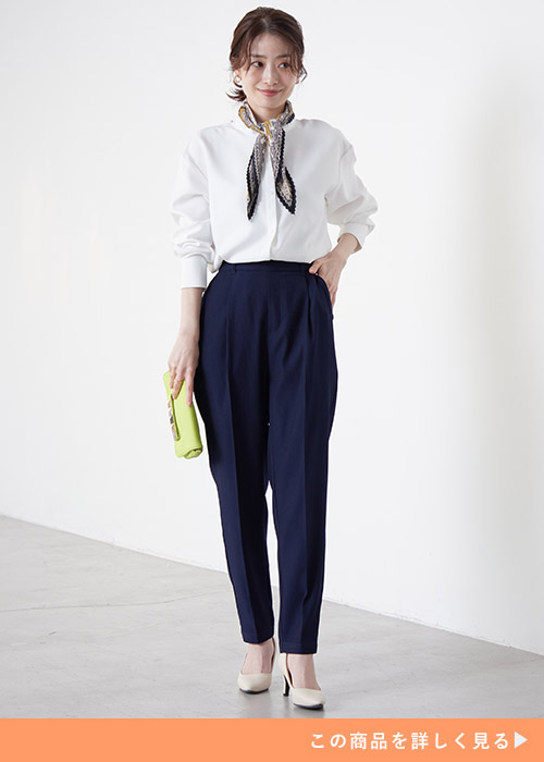 白ブラウスを着て、ネイビーのテーパードパンツを履き、スカーフを首に巻く女性
