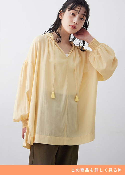 黄色のシアー素材ブラウスを着て、ブラウンのパンツを履く女性