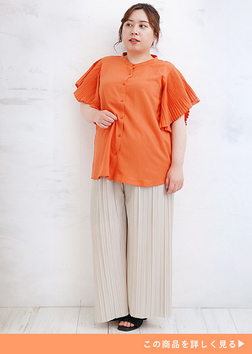 袖プリーツのオレンジトップスを着て、アイボリーのプリーツパンツを履く女性