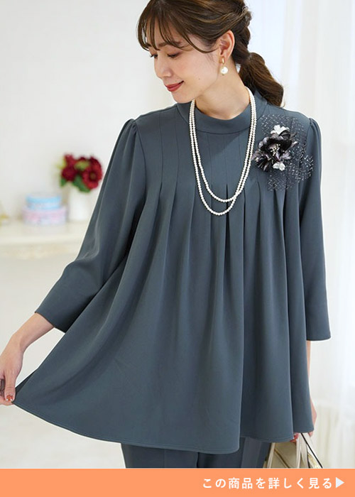 グレーのフレアチュニックを着た左胸に、パールネックレスと黒のコサージュをつけている女性