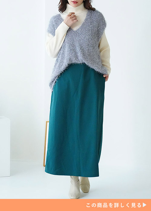 白のタートルネックニットにライトブルーのVネックベストを重ね着し、グリーンのタイトスカートを履く女性