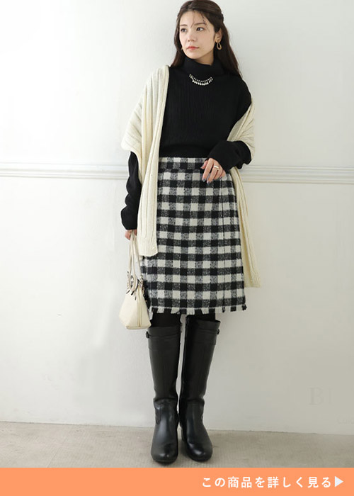 黒のタートルネックニット×チェック柄ミニスカートに白のジレを羽織り、ロングブーツを履く女性