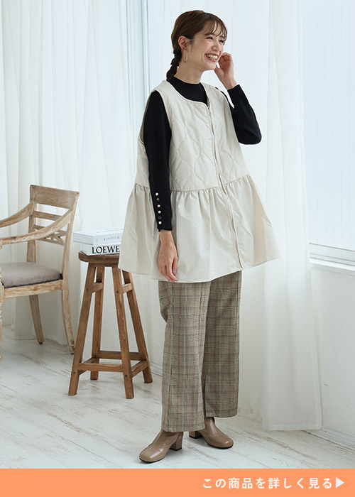 オフホワイトのひょうたんキルトベストに黒ニットトップスとブラウンチェックパンツを合わせる女性