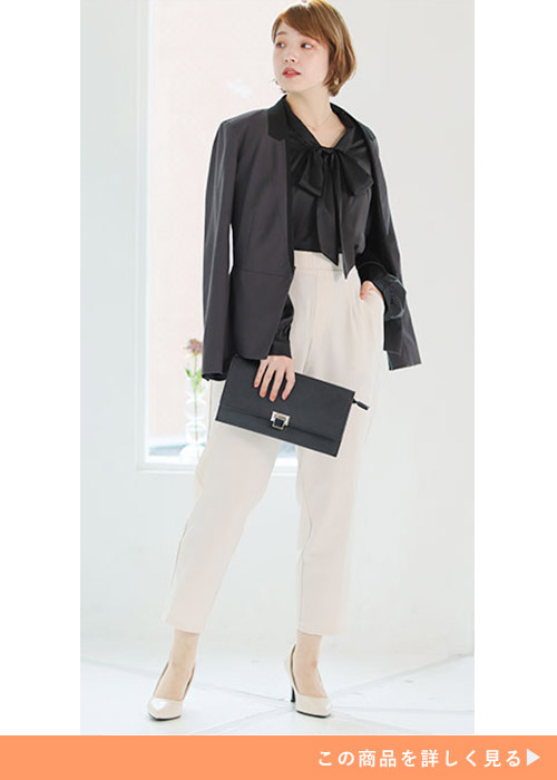 サテン素材のリボン付きのブラックブラウスにホワイトのパンツとパンプスを合わせる女性