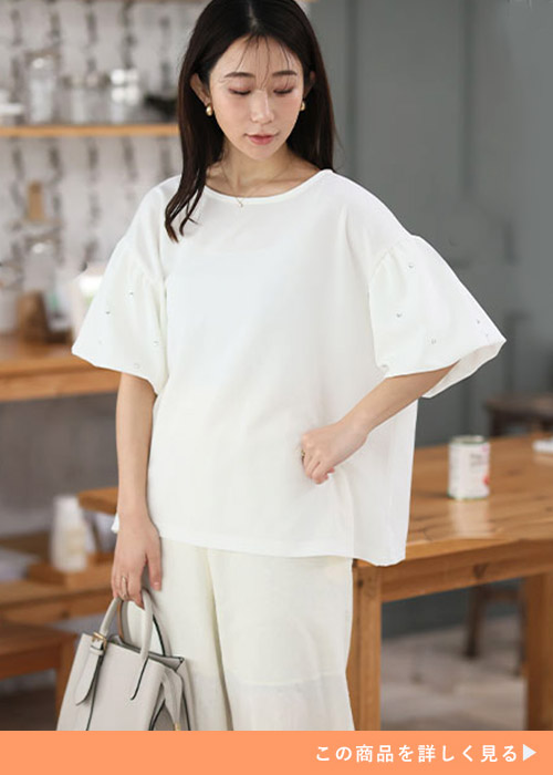 オフホワイトのビジュー付きプルオーバーとホワイトパンツのコーデにハンドバッグを持つ女性