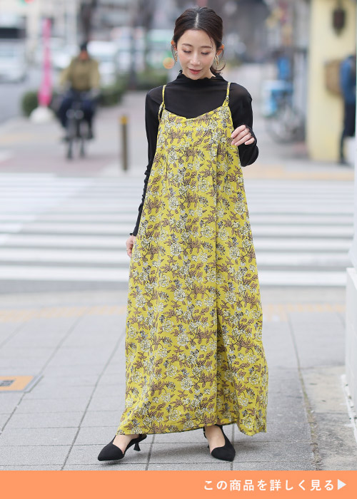 イエローの花柄ワンピースとブラックハイネックニットのレイヤードコーデにブラックパンプスを履く女性