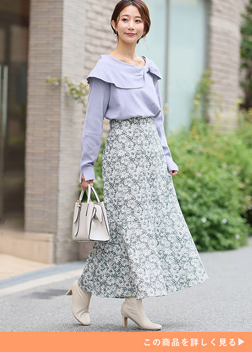 ホワイトの花柄スカートとパープルのリボントップスのコーデにレザーバッグとブーツを合わせる女性